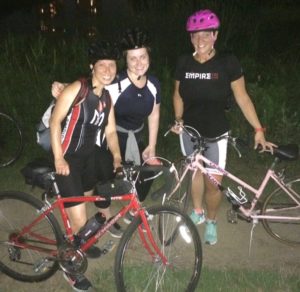 Three women posing with bicycles at night.